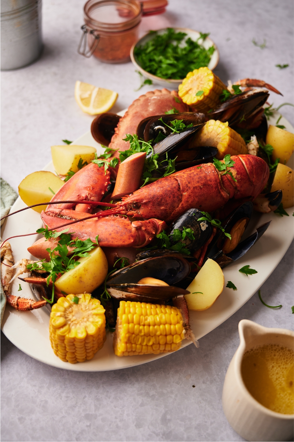 A large white serving tray piled with cooked clams, crab, lobster, corn, potatoes, sausage, and fresh herbs.