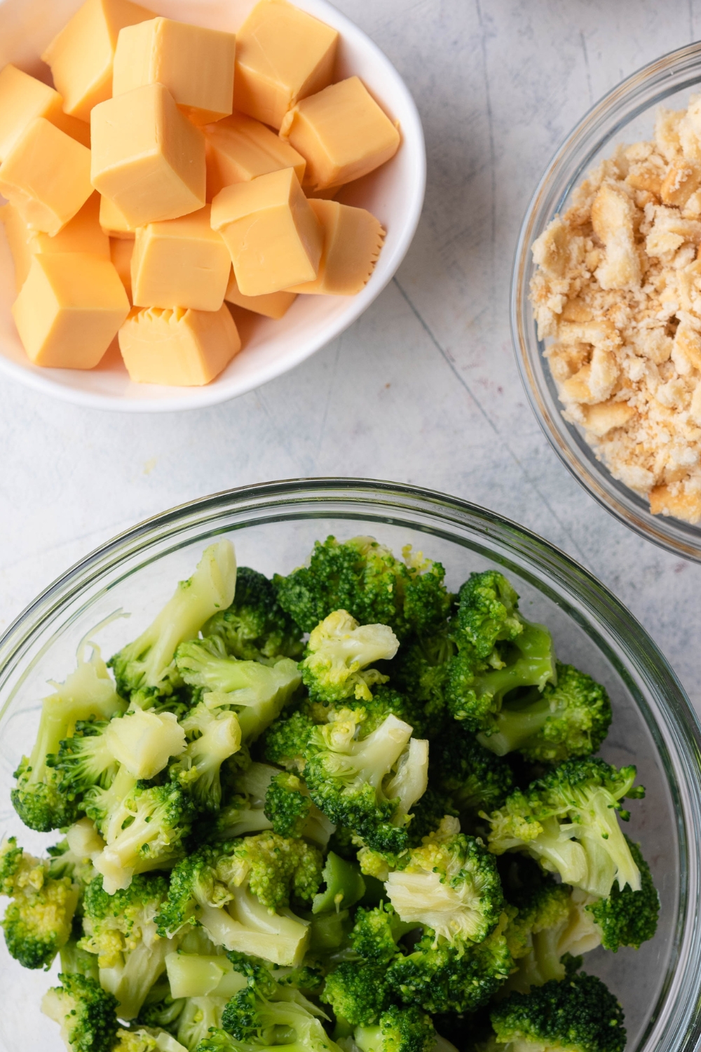 A close-up of three bowls containing broccoli, crumbled crackers, cubed cheese.