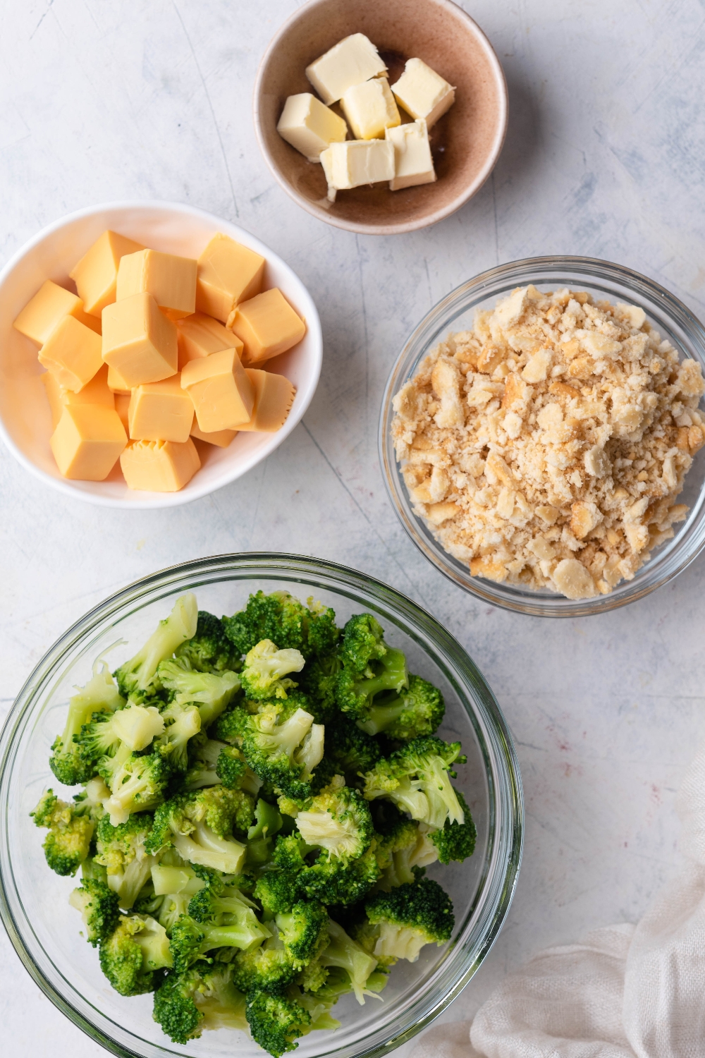 A countertop with multiple bowls containing broccoli, crushed crackers, cubed cheese, and cubed butter.