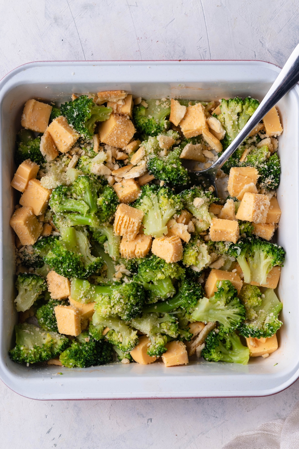 An overhead view of a casserole dish with broccoli, cubed cheese, and crumbled crackers mixed together.