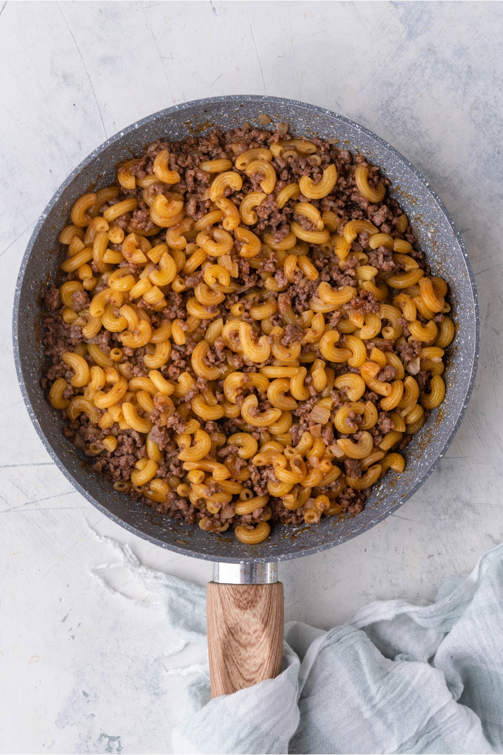 A skillet with cooked elbow macaroni and ground beef mixed together. There is a dish towel wrapped around the handle of the skillet.