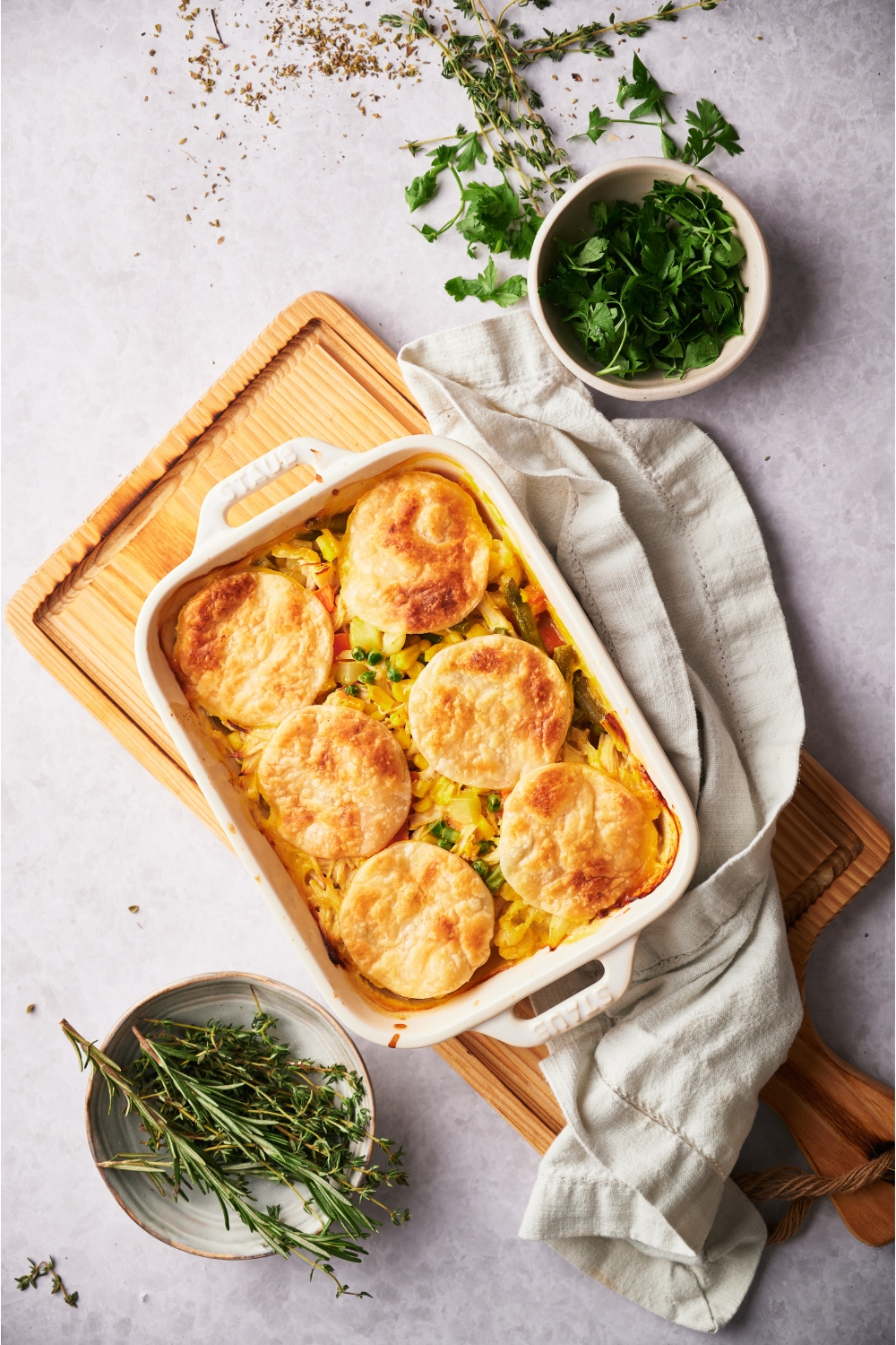 A white casserole dish with chicken pot pie casserole freshly baked. The dish is on a wood board and surrounded by bowls of fresh parsley and rosemary, as well as a dish towel.