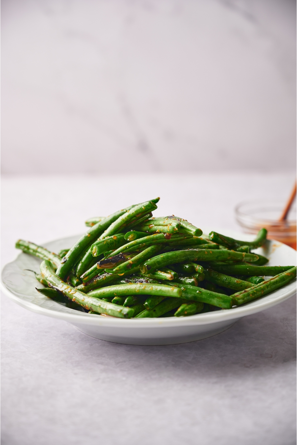 A white bowl filled with cooked and seasoned green beans. There is a bowl of honey in the background.