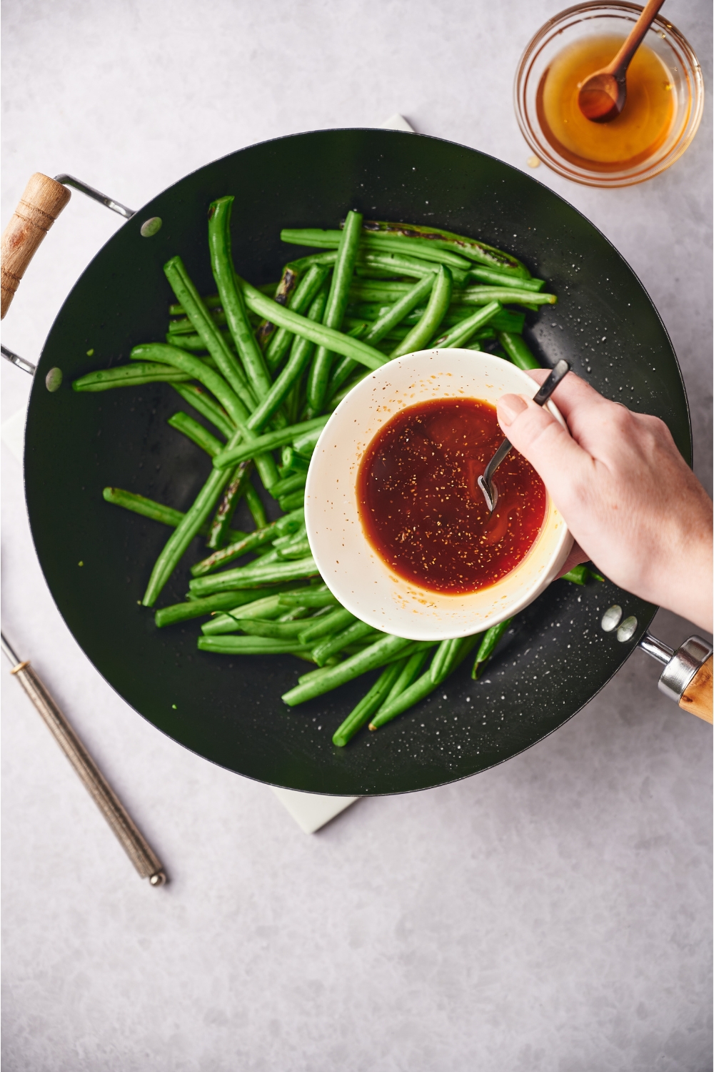 Black skillet with green beans and a hand holding a white ramekin above the skillet. The ramekin has a brown sauce. Next to the skillet is a small bowl of honey.
