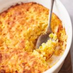 A casserole dish with corn souffle, a spoon is taking a scoop out of it.