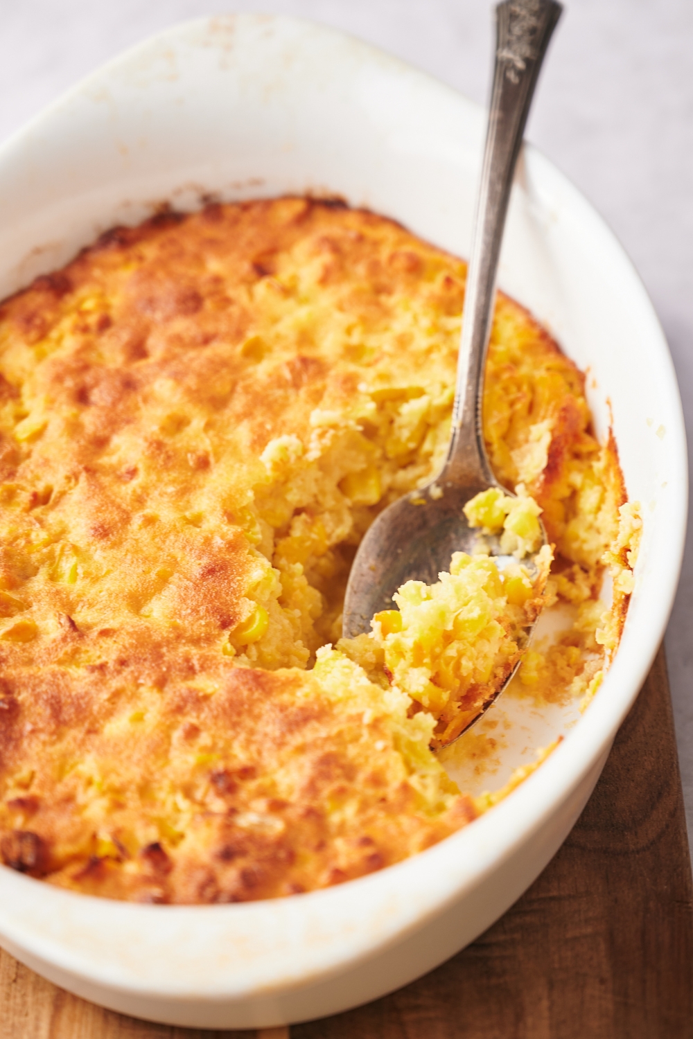 A casserole dish with corn souffle, a spoon is taking a scoop out of it.