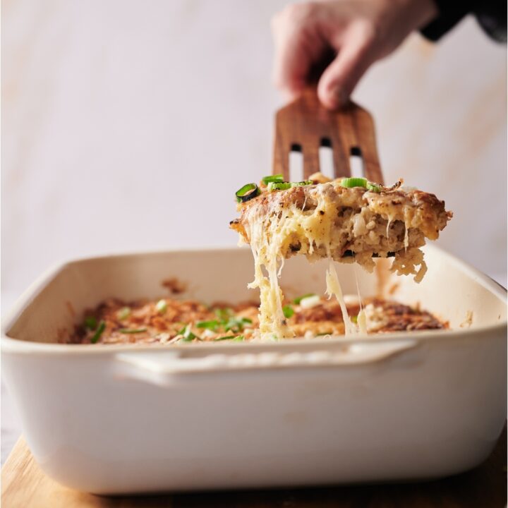 Hand scooping a serving of casserole out of a white casserole dish that is on top of a wood board.