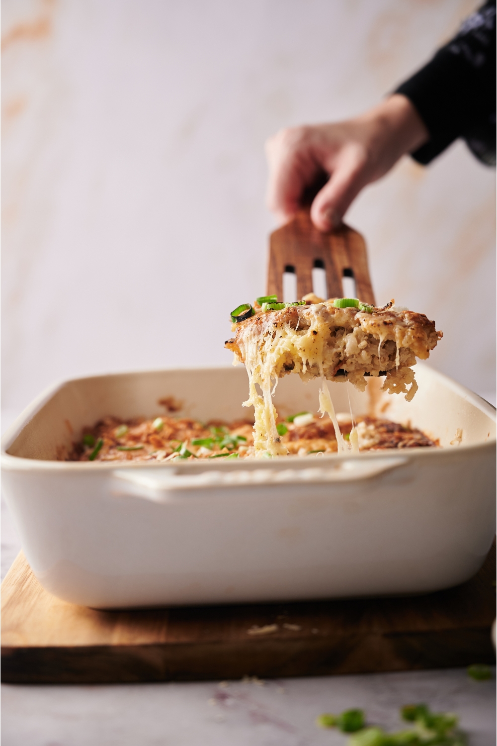 Hand scooping a serving of casserole out of a white casserole dish that is on top of a wood board.