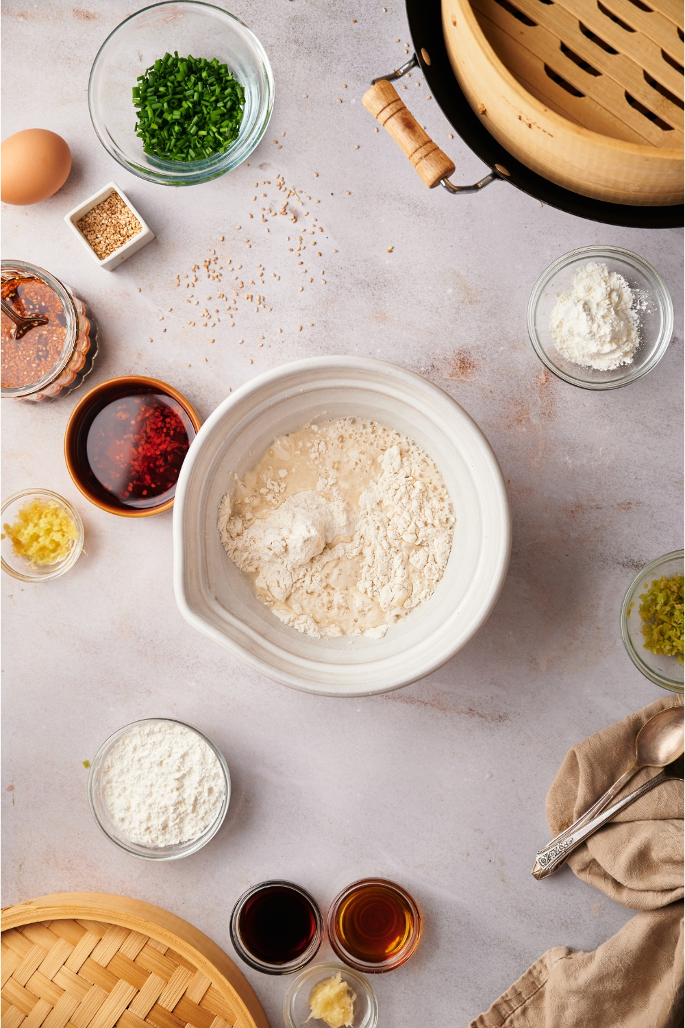 A white mixing bowl with a flour mixture, surrounded by bowls of ingredients including chili oil, garlic, sesame oil, and sesame seeds.