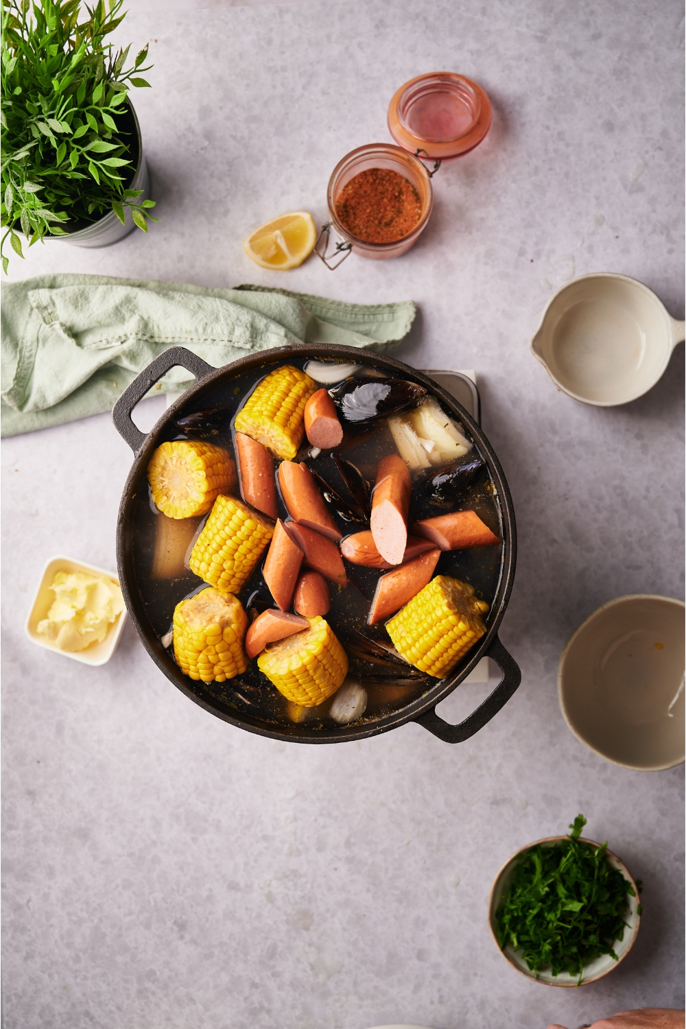 A black Dutch oven filled with sausages, clams and corn-on-the-cob pieces in a broth. Surrounding the Dutch oven is an assortment of ingredients including butter, fresh herbs, and spices.