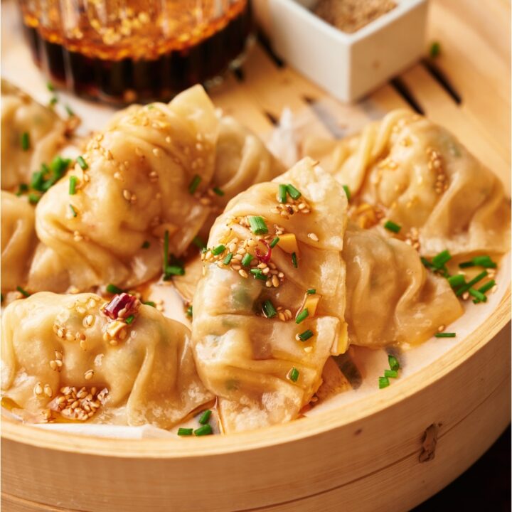 Steamed dumplings in a bamboo steamer on a wood board, garnished with green onion and sesame seeds.