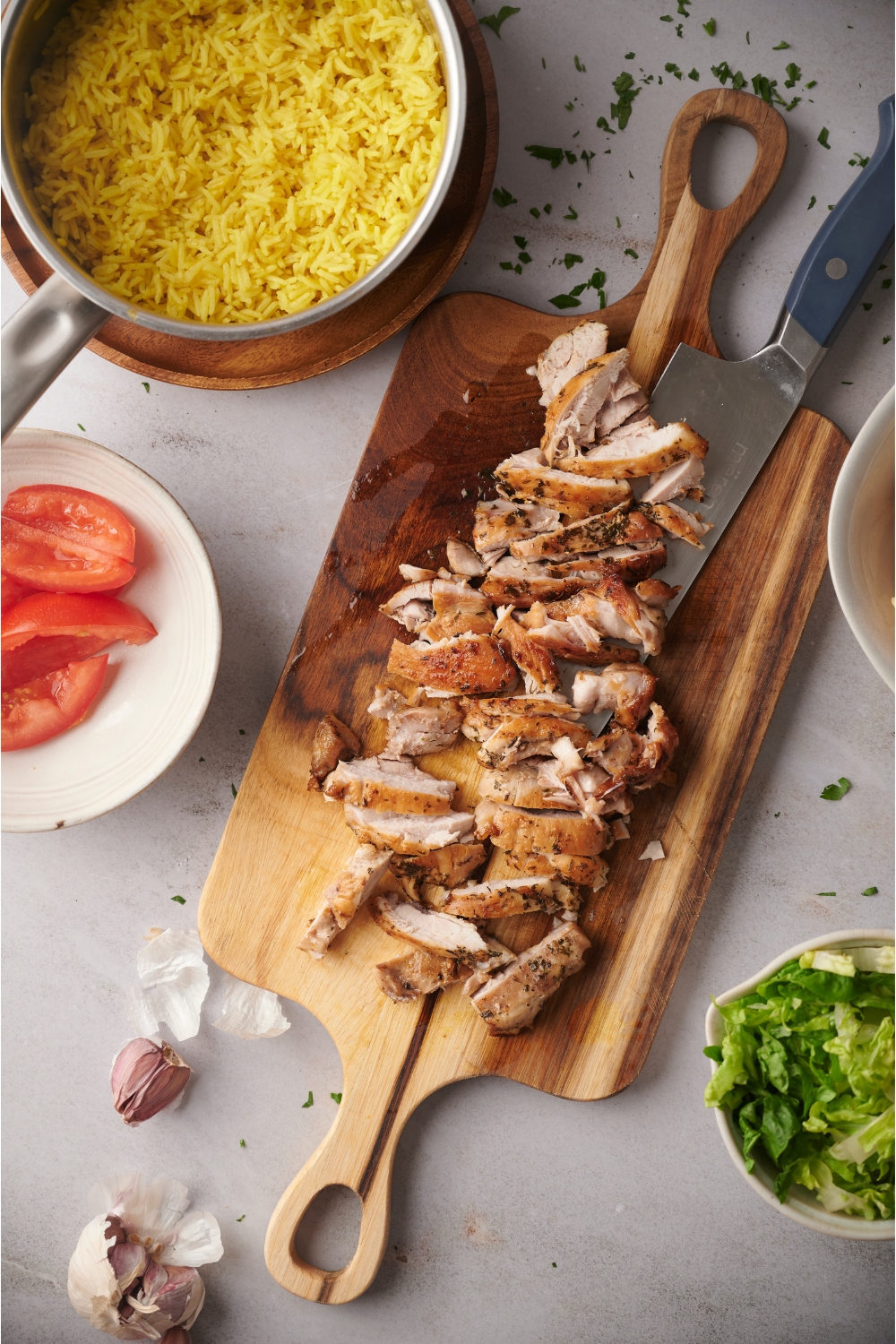 Seasoned chicken sliced thin on a wooden cutting board. There is a knife on the cutting board. Surrounding the chicken is an assortment of ingredients including a pot of yellow rice, tomatoes, garlic, and shredded lettuce.