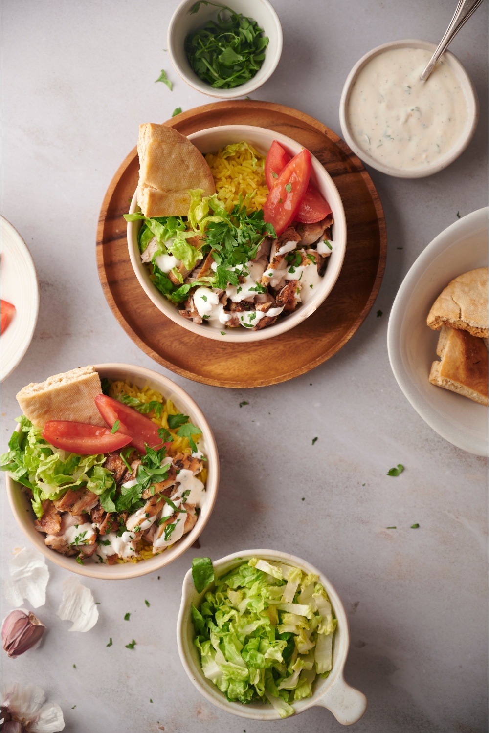 Bowls of halal chicken over rice, each with wedges of pita bread. One bowl is on a wooden plate. There is a small bowl of shredded lettuce and an assortment of other ingredients surrounding the bowls.
