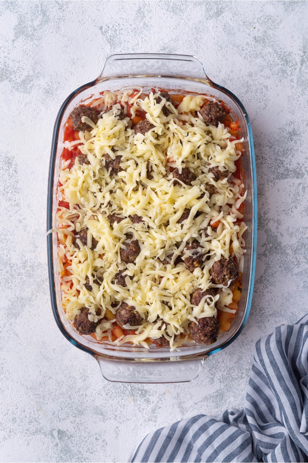 A clear baking dish filled with meatballs, pasta, red sauce, and shredded cheese.