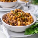 A white bowl filled with cheeseburger casserole that is garnished with fresh parsley. The bowl is on a white plate with a fork and parsley leaves on the plate as well. In the background there is a second bowl of cheeseburger casserole and raw ingredients for the casserole.