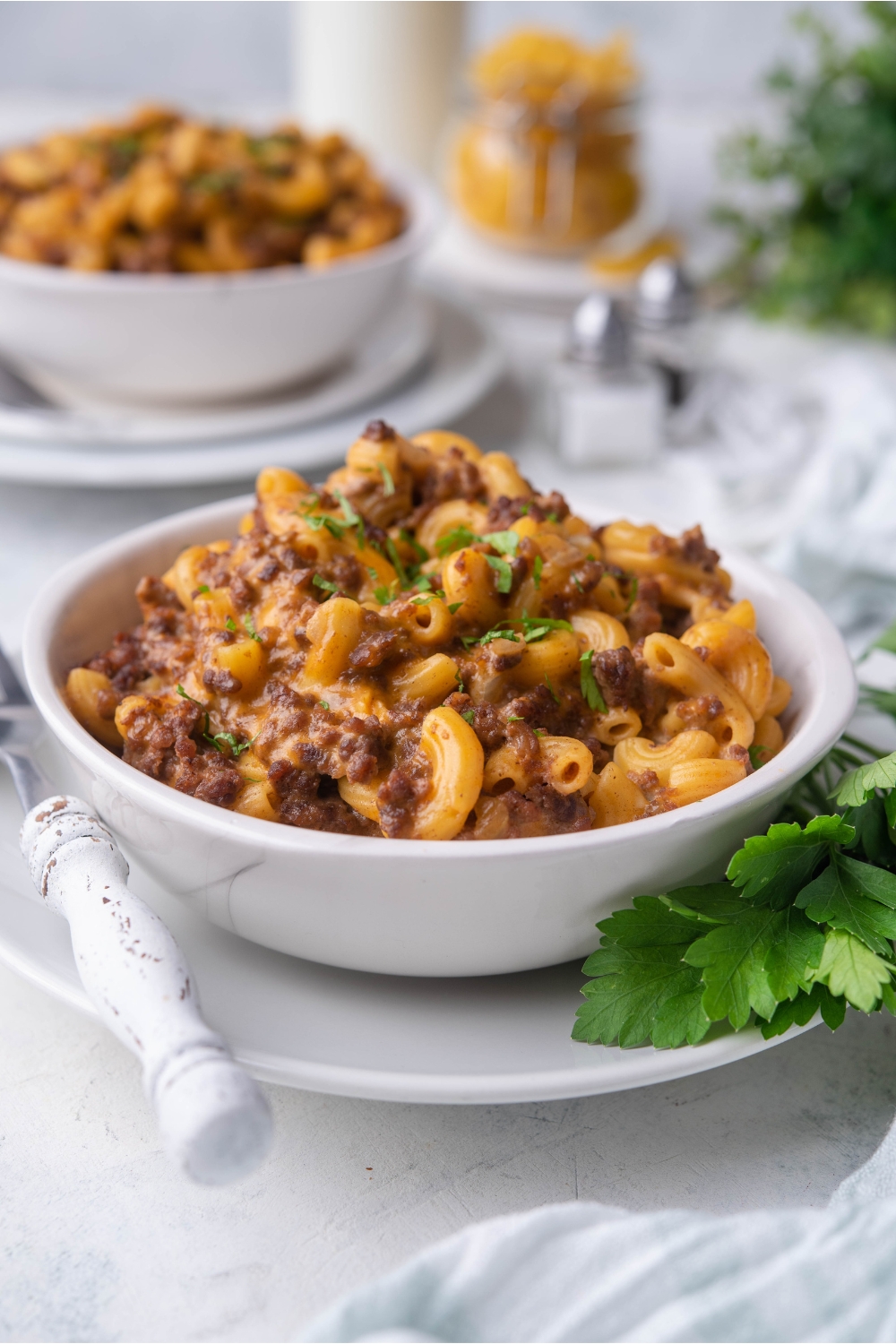 A white bowl filled with cheeseburger casserole that is garnished with fresh parsley. The bowl is on a white plate with a fork and parsley leaves on the plate as well. In the background there is a second bowl of cheeseburger casserole and raw ingredients for the casserole.