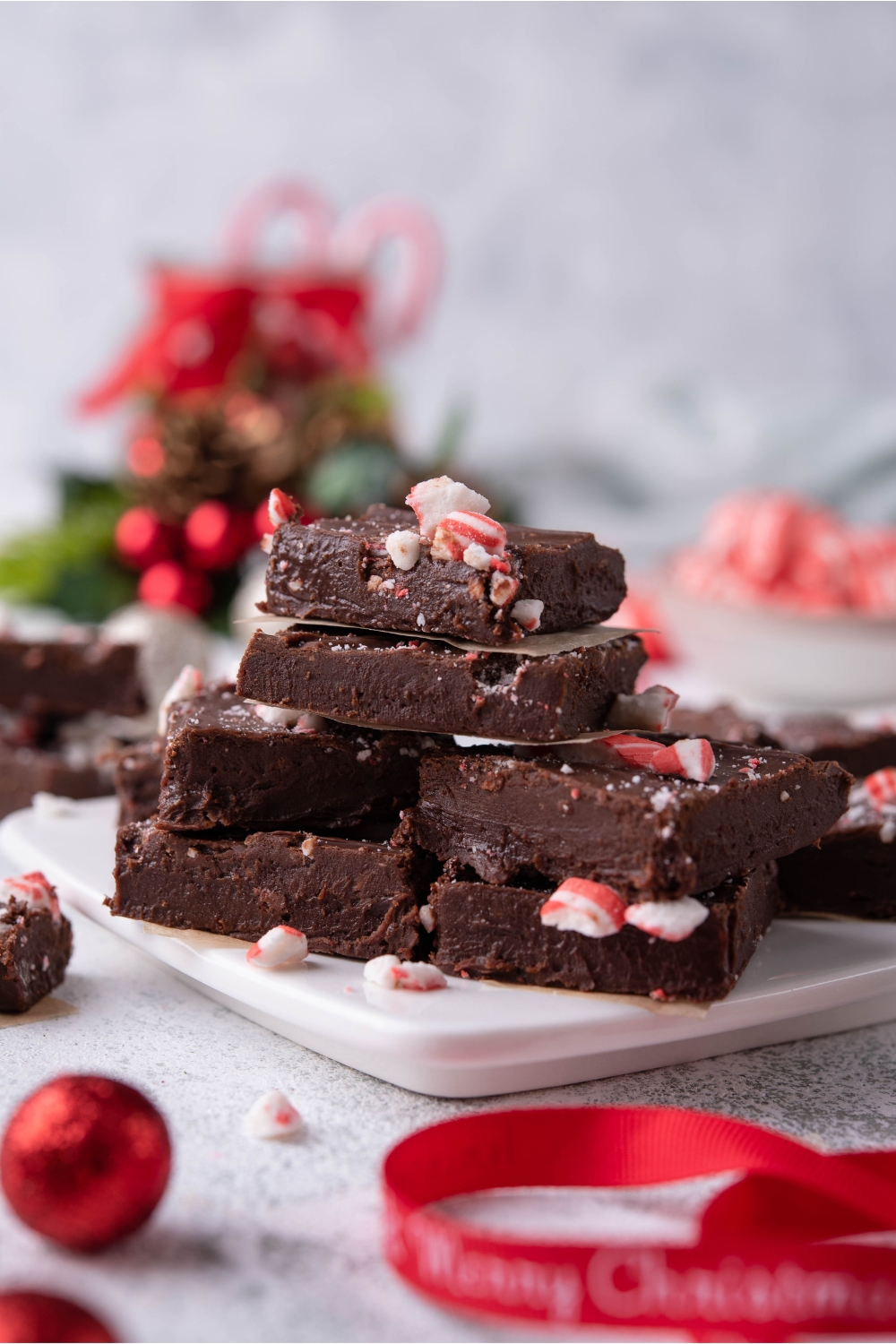 Squares of peppermint fudge stacked on top of one another on a white tray, with Christmas ornaments surrounding the fudge.