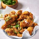 A blue bowl lined with parchment paper and filled with fried chicken pieces that have been garnished with fresh herbs. There is a bowl of fresh herbs in the background.