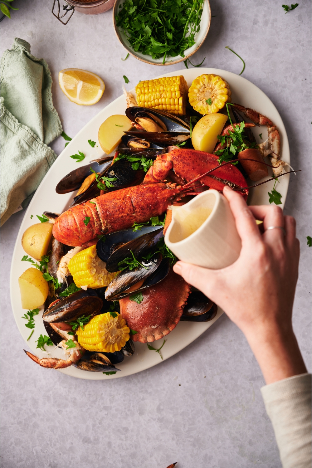 A hand pouring melted butter over a large white serving tray piled with cooked clams, crab, lobster, corn, potatoes, sausage, and fresh herbs.