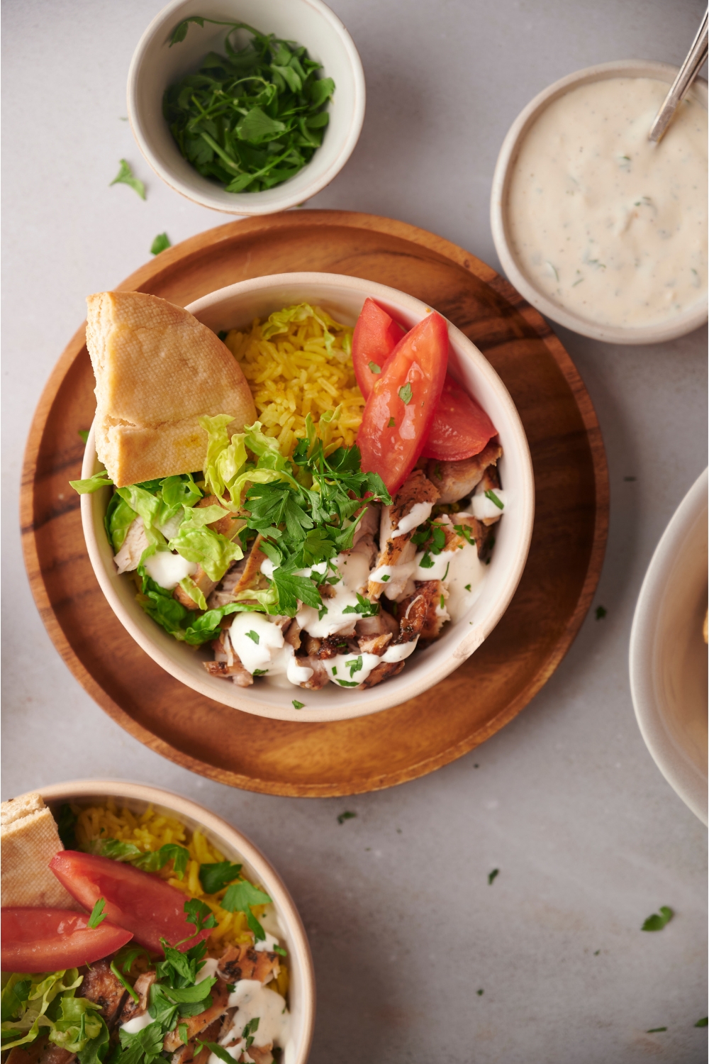 Halal chicken over rice in a white bowl on a wood plate. There is a wedge of pita bread in the bowl and an assortment of ingredients surrounding the bowl of chicken and rice.
