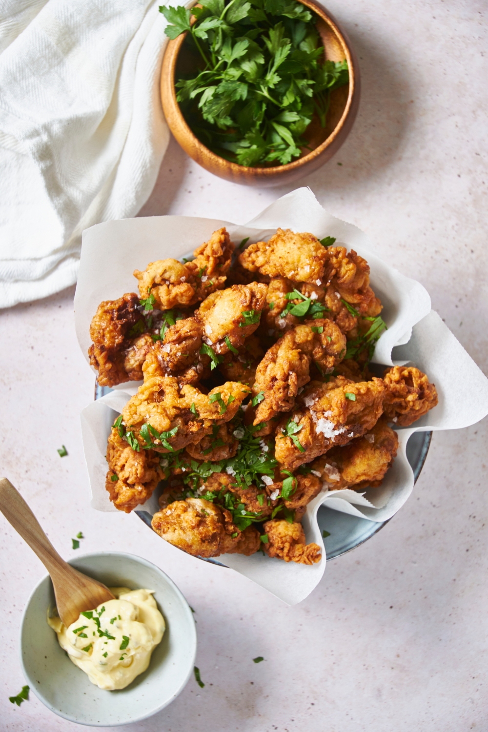 A bowl lined with parchment paper and filled with fried chicken that has been garnished with fresh herbs. Surrounding the bowl there is a white dish towel, a small bowl of fresh herbs, and a bowl with a spoonful of sauce in it.