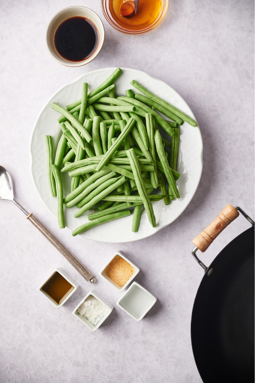 An assortment of ingredients including a plate filled with fresh green beans and small bowls of honey, soy sauce, and spices. There is a black skillet in the corner.