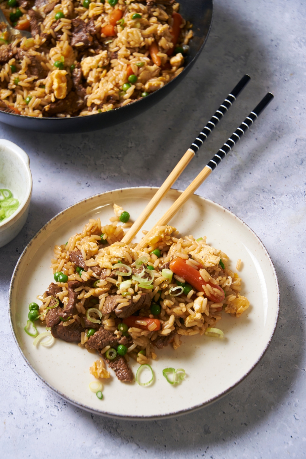 A white bowl with beef fried rice and a pair of chopsticks in the bowl. The rest of the fried rice is next to the white bowl.