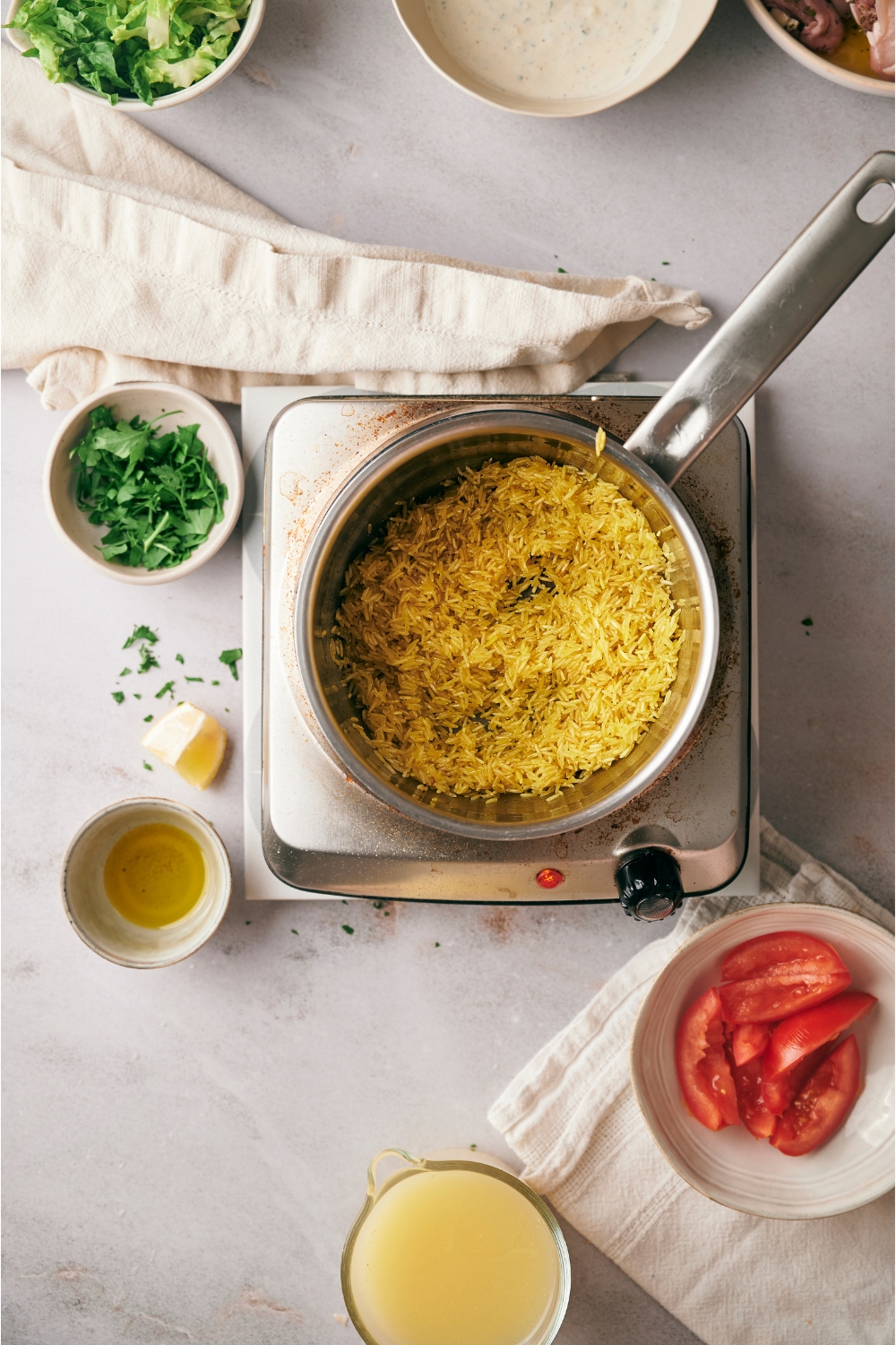 A saucepan over an electric burner. The saucepan is filled with yellow rice and is surrounded by an assortment of ingredients including tomato wedges, broth, butter, and parsley.