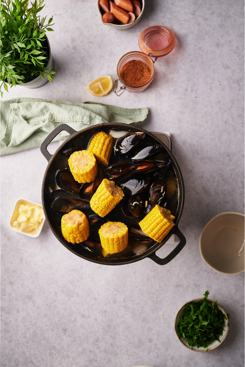 A black Dutch oven filled with clams and corn-on-the-cob pieces in a broth. Surrounding the Dutch oven is an assortment of ingredients including butter, fresh herbs, and spices.