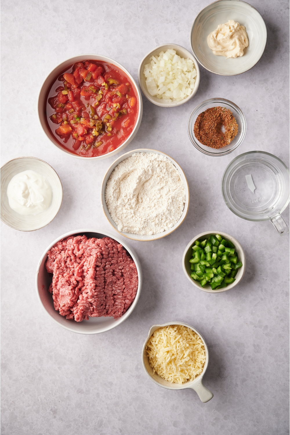 An assortment of ingredients including bowls of flour, sour cream, mayo, shredded cheese, salsa, raw ground beef, water, and seasoning.