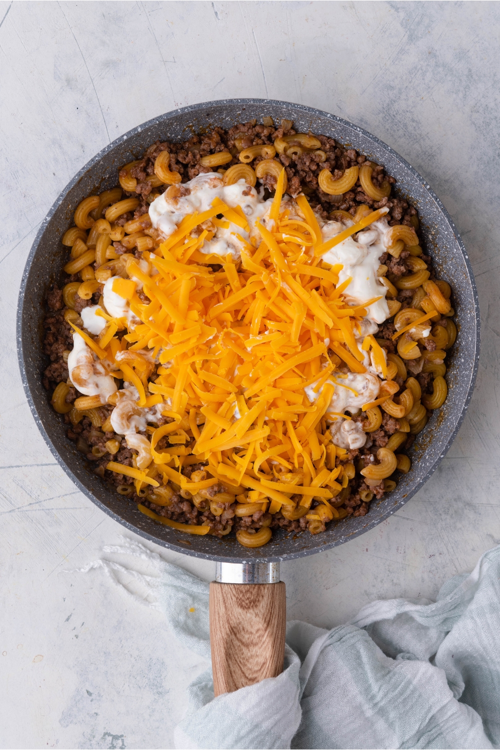 A skillet with elbow macaroni mixed in with ground beef. There is sour cream and shredded cheese topped directly in the center of the skillet and not yet mixed in. There is a dish towel wrapped around the handle of the skillet.