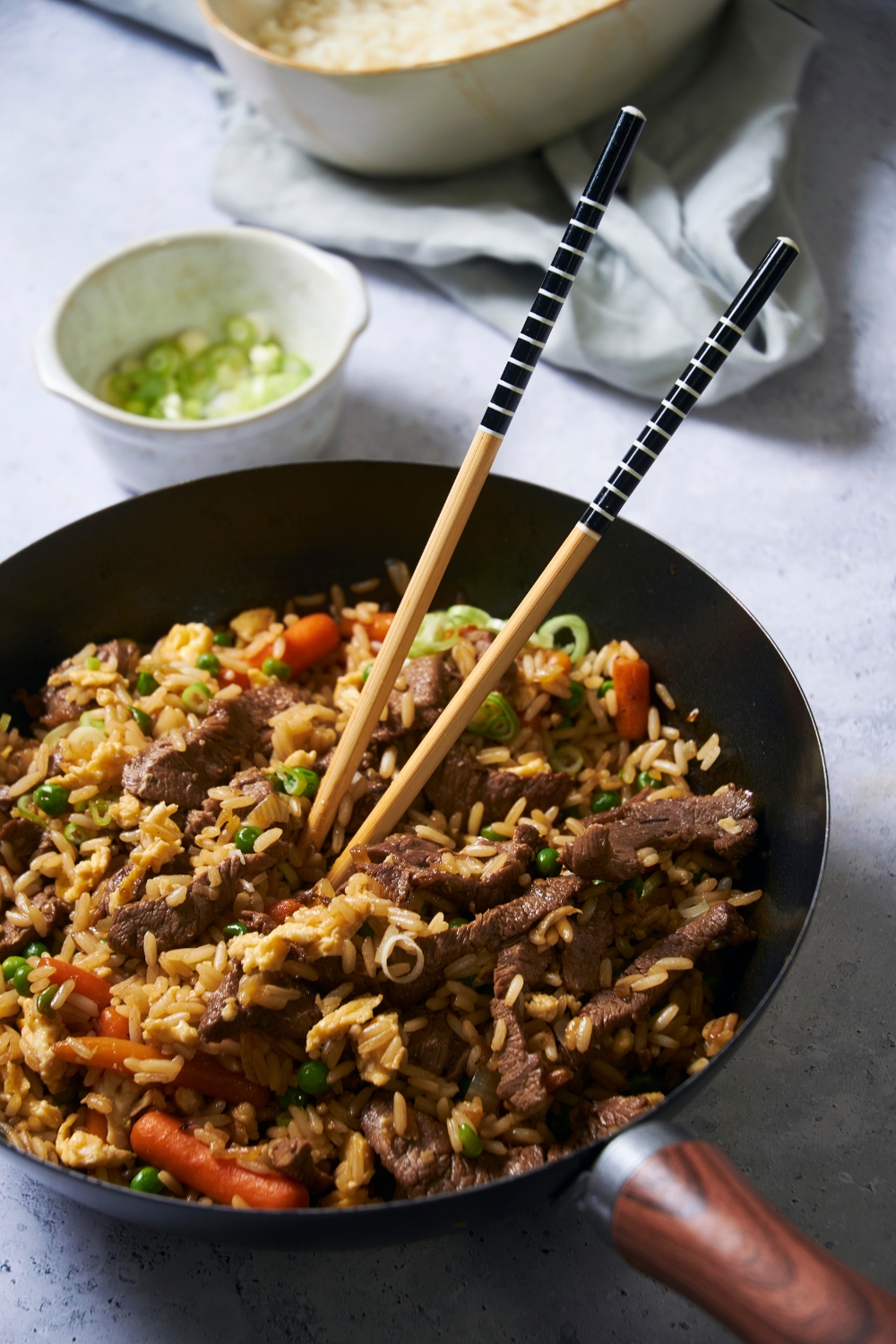 A black skillet filled with rice, peas, carrots, cooked beef, and green onions. There is a pair of chopsticks in the skillet and a small bowl of green onions in the background.