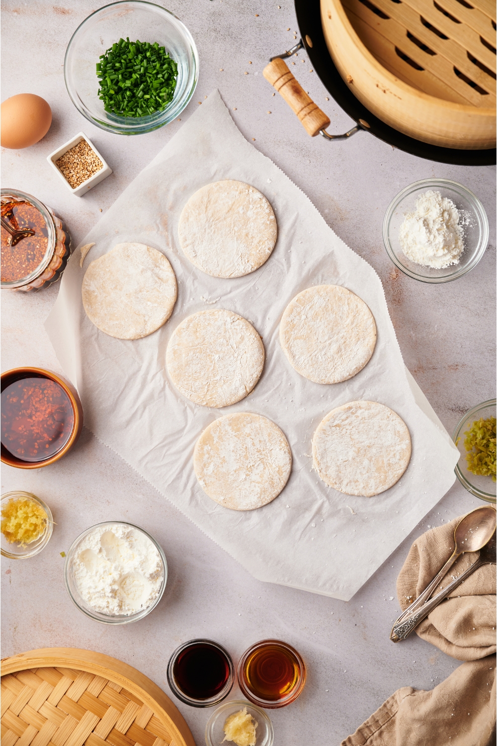 Six dough discs on a piece of white parchment paper, surrounded by an assortment of ingredients including bowls of oil, soy sauce and various dry ingredients.