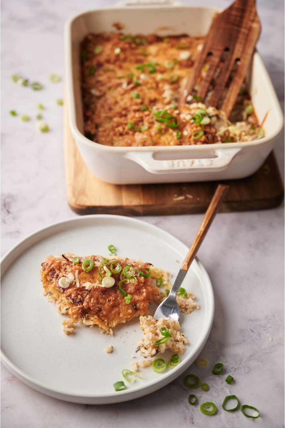 A white plate with a serving of pork chop casserole and a fork on the plate. The rest of the casserole is in a baking dish next to the plate.