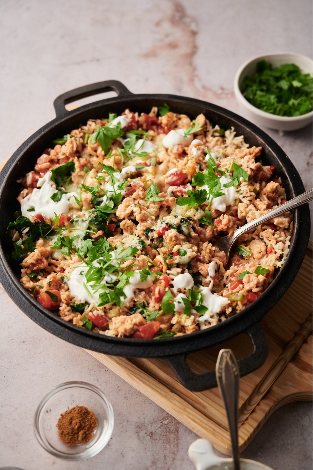 Black skillet on a wood board, filled with stuffed pepper casserole. There is a spoon in the pot and the casserole is topped with sour cream and fresh herbs.