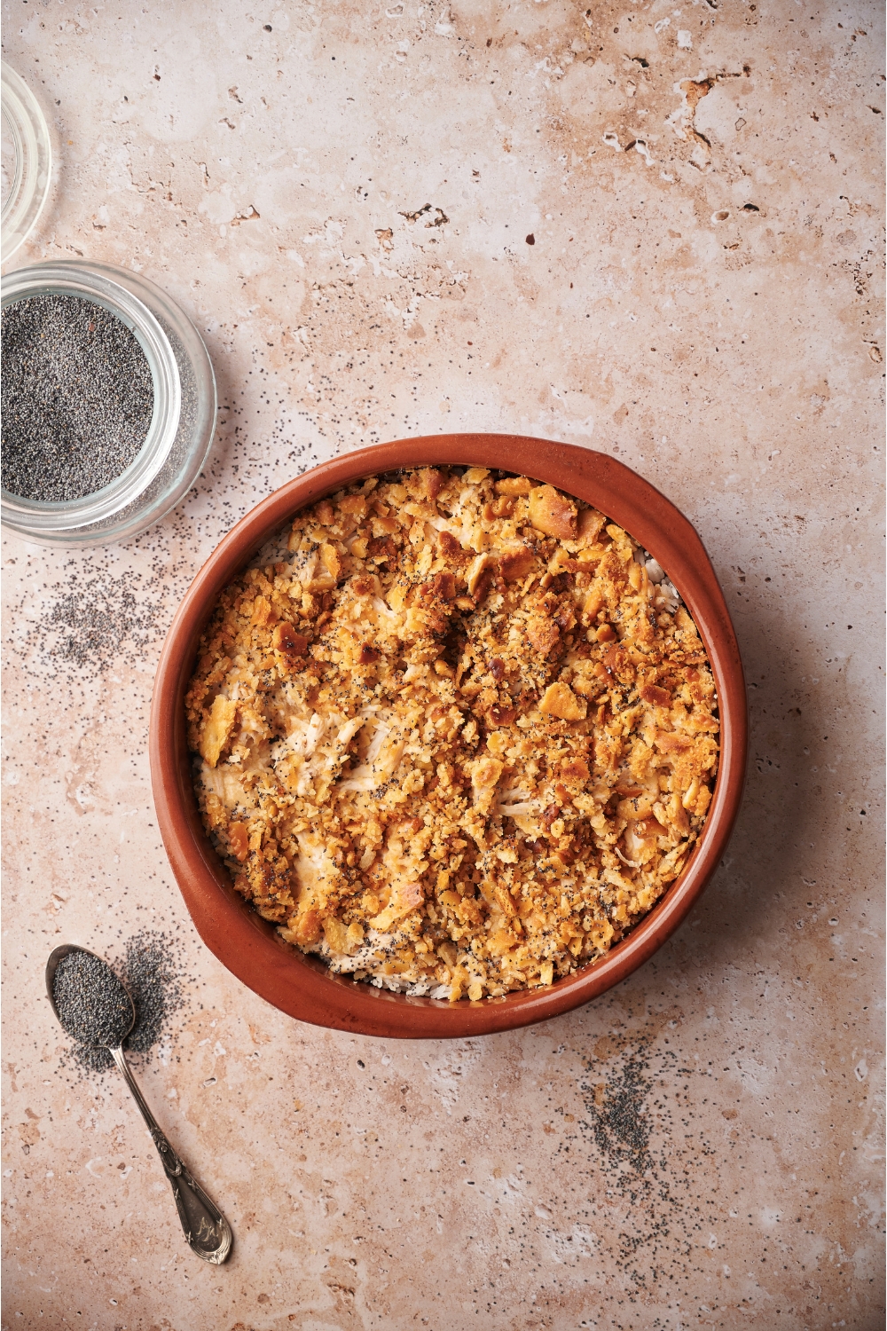 A red casserole dish filled with cooked chicken casserole. Next to the dish is a spoonful of poppy seeds and a bowl of poppy seeds.