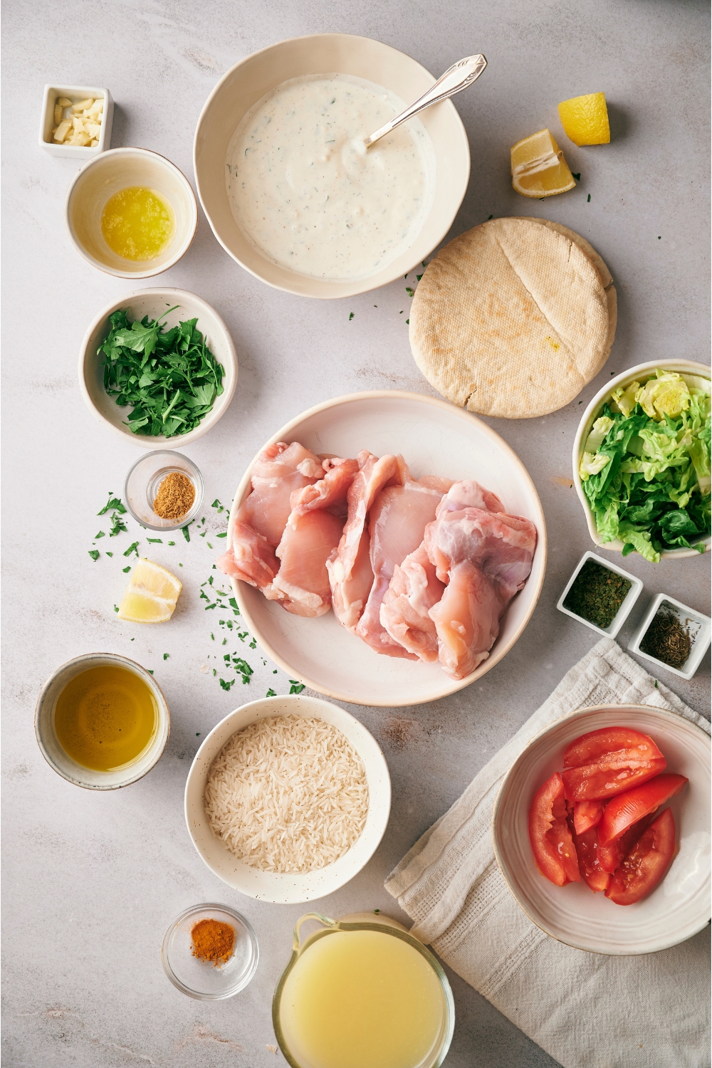 An assortment of ingredients for halal chicken over rice including bowls of raw chicken, white sauce, tomatoes, uncooked rice, broth, parsley, and pita bread.