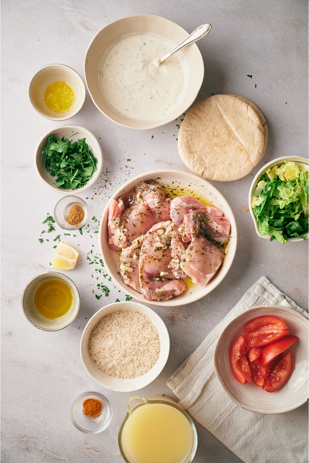 An assortment of ingredients for halal chicken over rice including bowls of raw marinating chicken, white sauce, tomatoes, uncooked rice, broth, parsley, and pita bread.