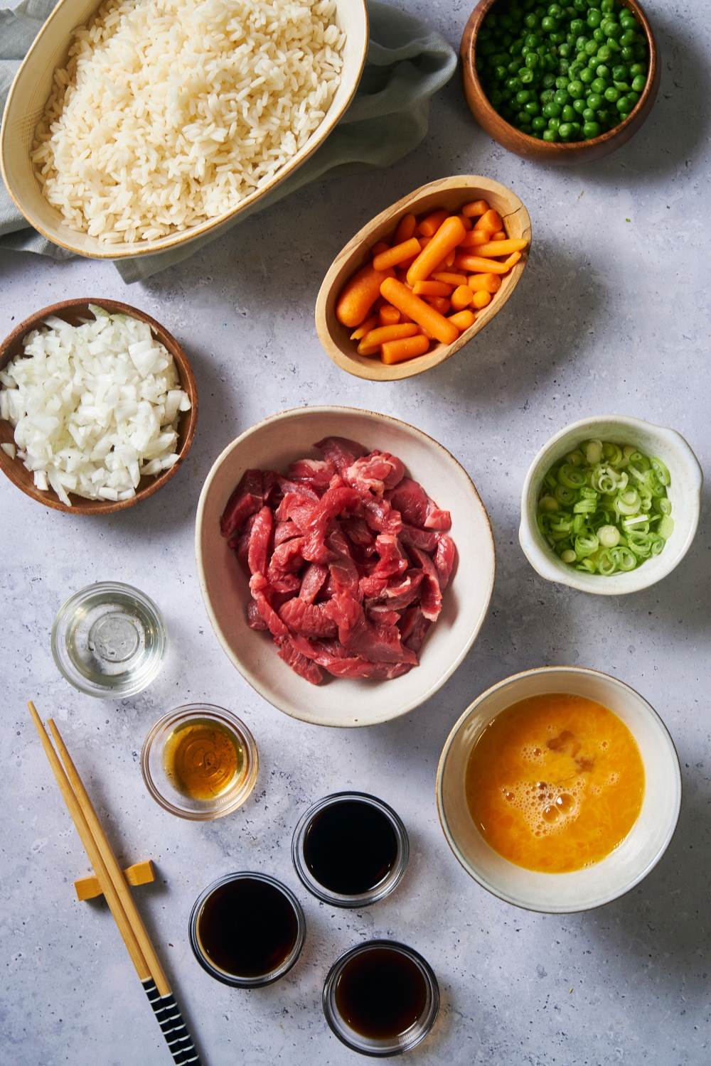 An assortment of ingredients including bowls of raw beef, carrots, peas, scallions, beaten eggs, onion, rice, and various sauces.