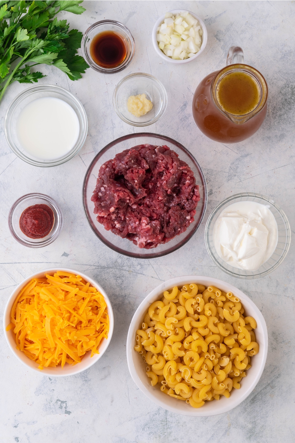 An assortment of ingredients for cheeseburger casserole including bowls of shredded cheese, uncooked elbow macaroni, raw ground beef, beef broth, sour cream, seasonings, diced onion, and tomato paste.