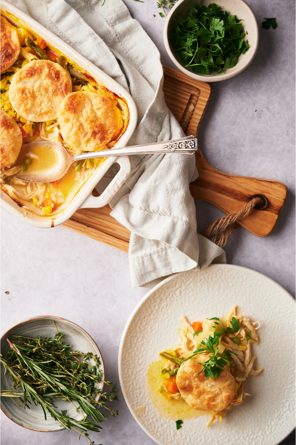 A white casserole dish with chicken pot pie casserole and a serving spoon in the dish. A serving of casserole has been portioned onto a white plate. The plate is garnished with fresh parsley. The casserole dish is on a wood board and next to it is a dish towel and bowls of fresh herbs.