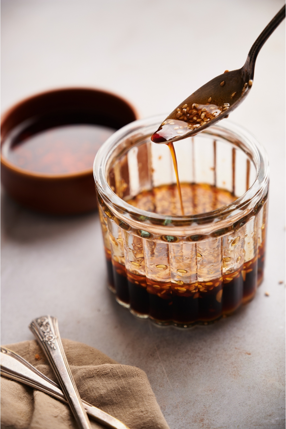 A spoon is dripping sauce into a clear jar. In the background there is a bowl of chili oil.