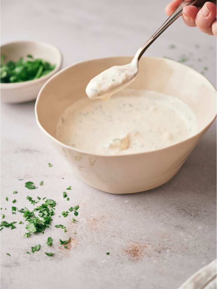 A hand holding a spoonful of halal white sauce. There is parsley sprinkled around the counter next to the bowl, and a bowl of parsley in the background.