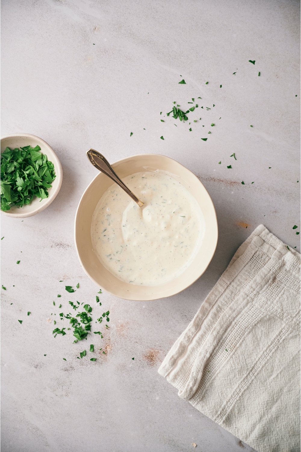 A white bowl filled with halal white sauce and a spoon in the bowl. There is a bowl of parsley and a kitchen towel next to the bowl.