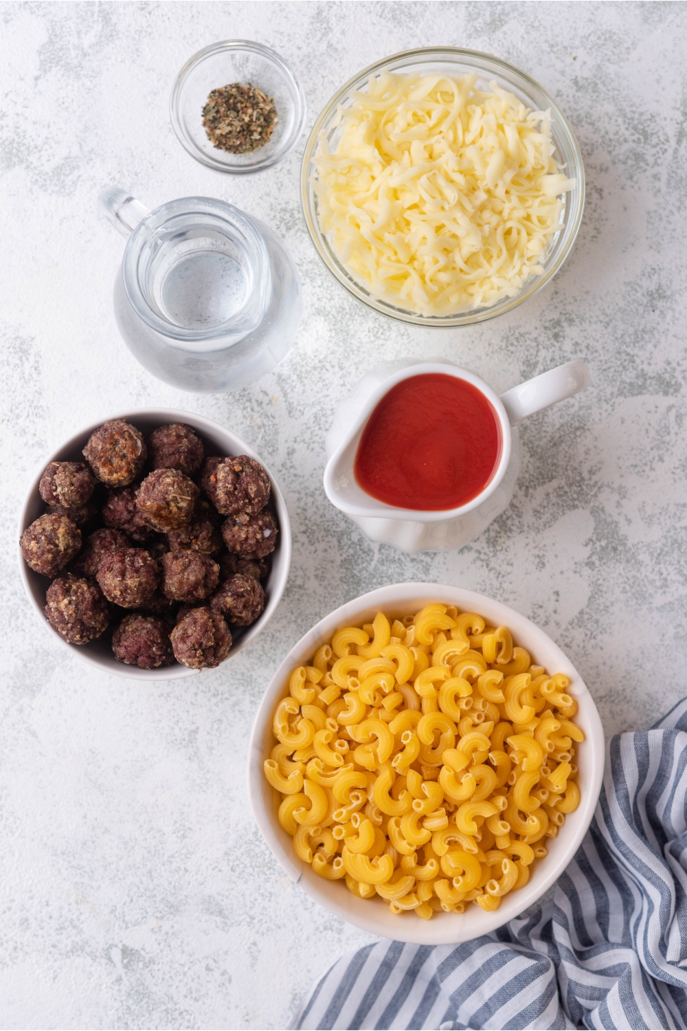 An assortment of ingredients including bowls of meatballs, elbow macaroni, shredded cheese, water, seasoning, and tomato sauce.