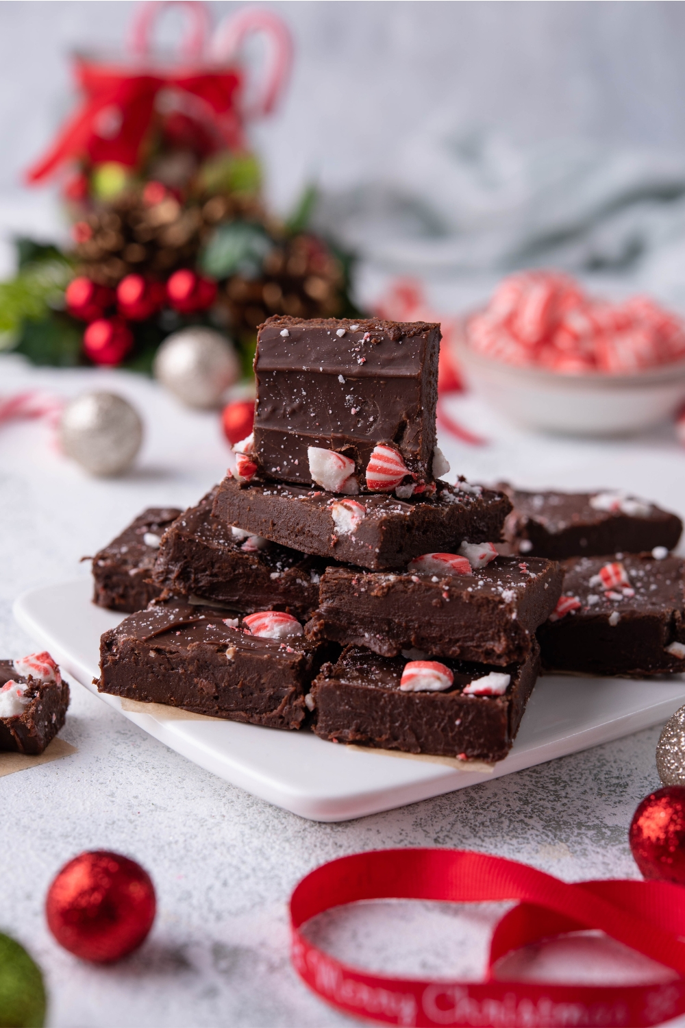 Squares of peppermint fudge stacked on top of one another on a white tray, with Christmas ornaments surrounding the fudge.