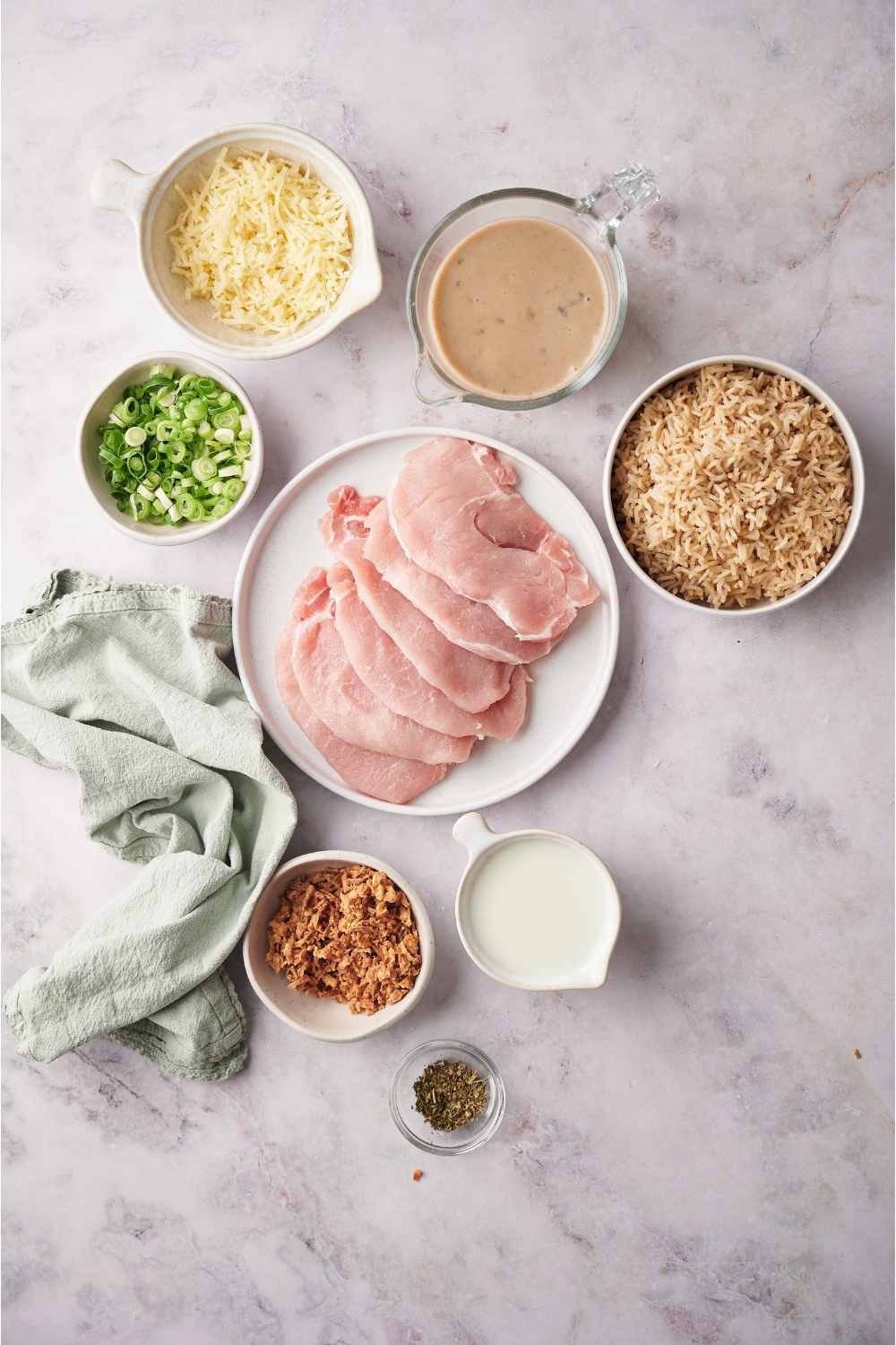 An assortment of ingredients including a plate of raw pork chops and bowls of cooked rice, soup, shredded cheese, green onion, sour cream, and seasonings.