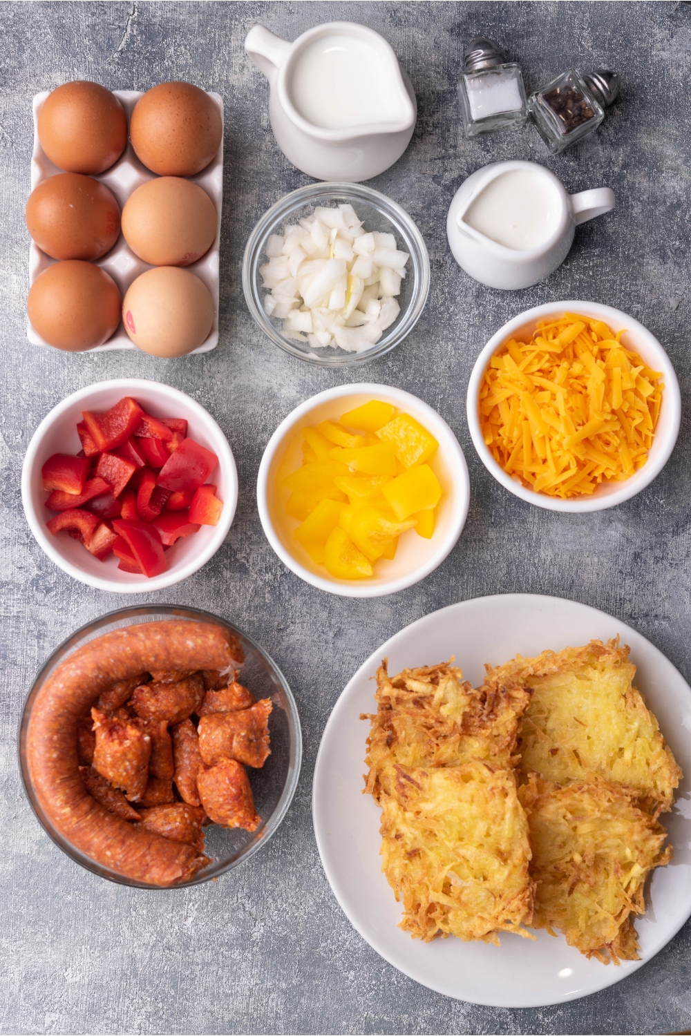 An assortment of ingredients for sausage casserole including a bowl of uncased sausage, bowls of bell peppers, shredded cheese, diced onion, a plate of hash browns, a half dozen eggs, cream, milk, and salt and pepper shakers.