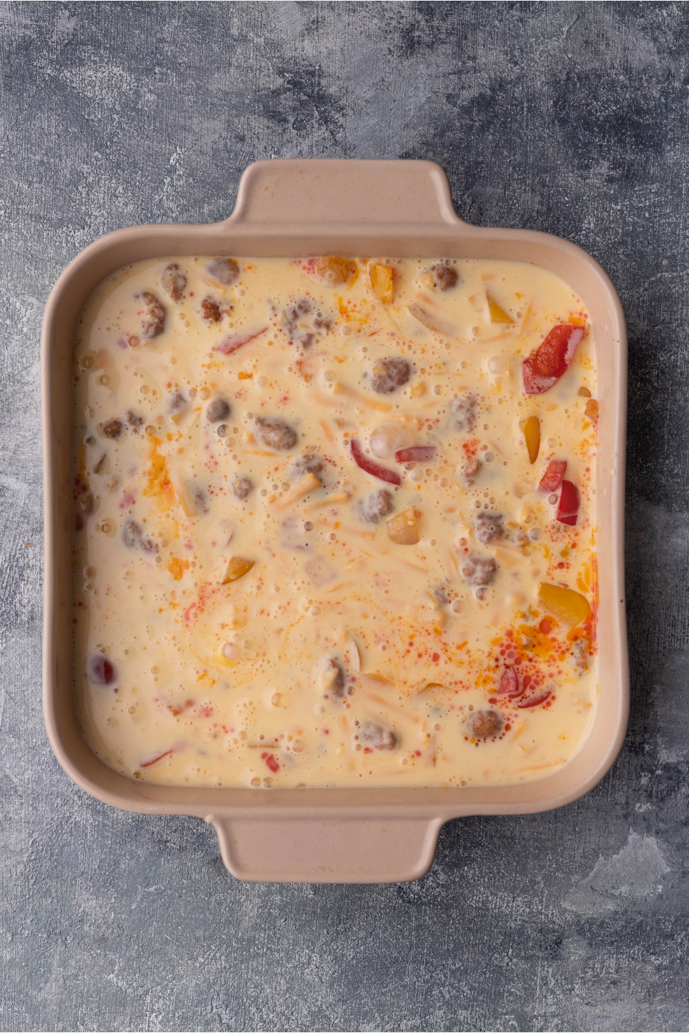 A brown casserole dish with sausage, peppers, onions, and an egg and cream mixture has been added to the dish. The dish has not yet been baked and is still liquidy.