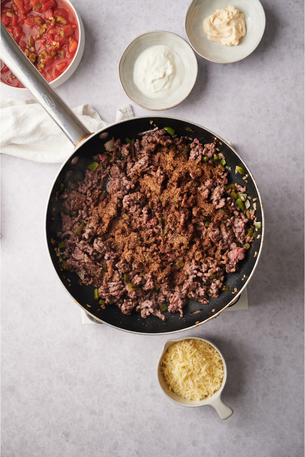 A black skillet filled with ground beef, peppers, onions, and seasoning. Surrounding the skillet are bowls of shredded cheese, sour cream, mayo and salsa.